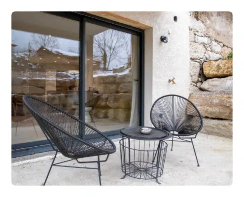 Location Keasy - The Barn premium Un patio accueillant avec deux chaises en osier noir et une table ronde posée sur un sol en pierre. Le reflet des arbres dans les portes vitrées suggère une atmosphère sereine et rustique.