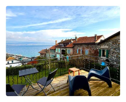 Location Keasy - Studio Lac terrasse vue lac Vue panoramique depuis une terrasse en bois avec chaises et table modernes, donnant sur des maisons en pierre aux toits rouges, se détachant sur un lac serein et un ciel nuageux.