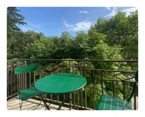Location keasy - Le Cocooning balcon Une table de bistrot verte et deux chaises sur un balcon, entourées d'arbres luxuriants sous un ciel bleu, créent un cadre extérieur paisible et accueillant.