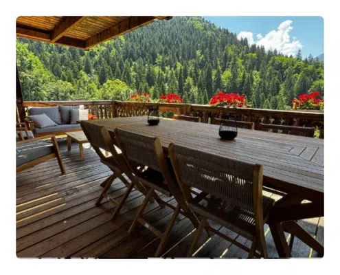 Location keasy - Le refuge vue panoramique · dépaysement total Une terrasse en bois surplombant des montagnes verdoyantes, aménagée avec une longue table et des chaises. Des fleurs rouges ornent la rambarde, créant une ambiance sereine et accueillante.